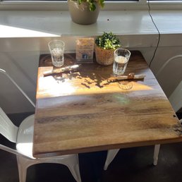 A wooden table positioned near a window with sunlight casting shadows on its surface. The table has two glasses of water, each with a napkin or cloth wrapped around the stem, and a small potted plant with green foliage. A business card or flyer is placed upright on the table. The window ledge behind the table holds another potted plant, and a black wire runs down from the window. Two white chairs are positioned on either side of the table, and the overall setting appears to be in a casual dining or cafe environment.