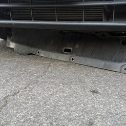 Improperly installed splash guard. - The image shows a black plastic splash guard beneath the front bumper of a vehicle, which appears to be improperly installed or damaged, as it is hanging down and not flush with the bumper. The vehicle is parked on a paved surface with visible cracks. The splash guard has multiple mounting holes and a central slot, indicating it is designed to be securely attached to the vehicle's underside.