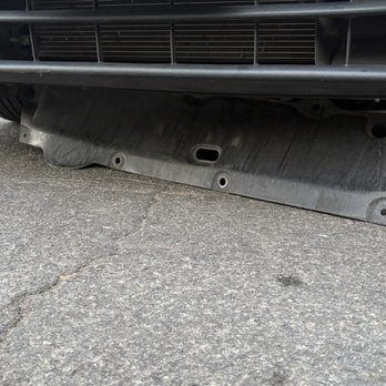 Honda North - Improperly installed splash guard. - The image shows a black plastic splash guard beneath the front bumper of a vehicle, which appears to be improperly installed or damaged, as it is hanging down and not flush with the bumper. The vehicle is parked on a paved surface with visible cracks. The splash guard has multiple mounting holes and a central slot, indicating it is designed to be securely attached to the vehicle's underside. - Auto Repair Near Me - Clovis, California