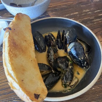 Harbinger - Mussels - A dish of mussels served in a creamy sauce in a black bowl, accompanied by a slice of toasted bread, on a wooden table. - New American Near Me - Des Moines, Iowa