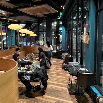 Geppetto - The image shows the interior of a restaurant with a modern design, including large floor-to-ceiling windows, wooden flooring, and ceiling panels with decorative patterns. Several diners are seated at tables, some wearing masks, and a server is attending to customers. The lighting includes hanging yellow lamps, and the space appears spacious with additional tables along the window wall. The overall atmosphere is contemporary and well-lit. - Italian Near Me - Cambridge, Massachusetts