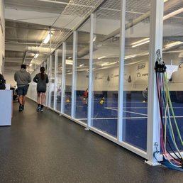 The scene from my waiting chair - The image shows an indoor sports facility featuring a glass-enclosed court, likely used for tennis or pickleball. The court has a blue surface with yellow balls and cones visible inside. The ceiling has exposed pipes and bright overhead lighting. Two people are walking along the corridor outside the court, and colorful resistance bands are hanging on the right side. The environment appears clean and well-maintained, suitable for recreational or training activities.