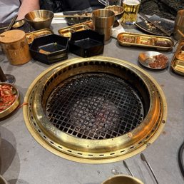 1 small grill for 8 people - The image shows a restaurant table with a built-in circular grill in the center, which is currently unused. Surrounding the grill are various dishes, bowls, and utensils, some with remaining food. There are glasses of beer, metal cups, and small plates on the table. The scene includes partially visible people seated around the table, indicating a social dining setting, likely at a Korean or Japanese barbecue restaurant.