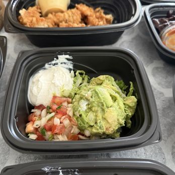Silverado - A black plastic food container holds a serving of guacamole, sour cream, and pico de gallo, with diced tomatoes, onions, and cilantro. Surrounding containers include fried chicken and other dishes, indicating a meal with Mexican or Tex-Mex cuisine. - American Near Me - Annandale, Virginia