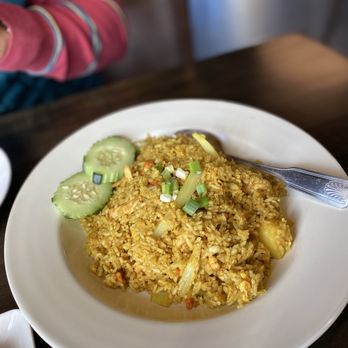 Dillon Thai restaurant - Pineapple Fried Rice - A white round plate contains pineapple fried rice with visible chunks of pineapple, rice, and vegetables, garnished with cucumber slices and chopped green onions. The dish is served on a dark wooden table, with a metal spoon resting on the edge of the plate. Part of a person's arm wearing a pink and blue striped sleeve is visible in the background, indicating a casual dining setting. - Thai Near Me - Dillon, Colorado