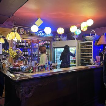 Chopin Theatre - The image shows a bar interior with a wooden counter and decorative lighting including colorful lanterns hanging from the ceiling. Two women are behind the bar, one reaching into a refrigerator stocked with drinks. The background features shelves with bottles and decorative items, creating a cozy, eclectic atmosphere. - Performing Arts Near Me - Chicago, Illinois