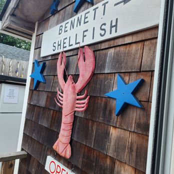 Bennett Shellfish - The image shows a wooden exterior wall with a sign for Bennett Shellfish, including a large lobster decoration hanging below the sign. The sign has the business name and an arrow pointing to the right. Blue star decorations are mounted on the wall around the sign. An open sign with hours is also visible, indicating the business is open from 11 to 8. The setting appears to be an outdoor area of a seafood business specializing in shellfish. - Seafood Near Me - Montauk, New York