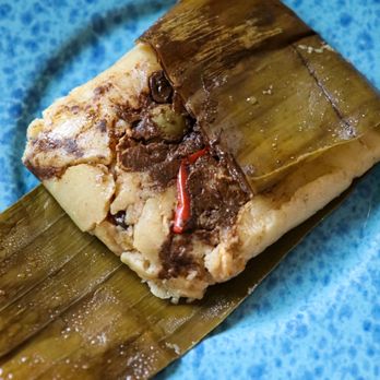 Morels - Chicken Tamale     Instagram.com/Maryelda - A chicken tamale wrapped in a banana leaf is placed on a blue ceramic plate. The tamale's filling includes chicken, red chili, and other ingredients, with some of the filling visible at the open end. The banana leaf wrapping is partially peeled back, revealing the tamale inside. The plate is set on a light-colored surface. - Food Trucks Near Me - Newport, Oregon