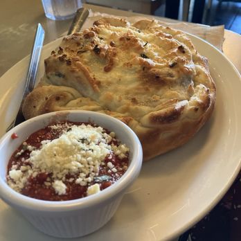 Pegasus Pizza - Pegasus Calzone - A baked calzone with a crispy, golden-brown crust is presented on a white plate, with visible toppings and a slightly uneven surface. Next to it, a small white bowl contains marinara sauce garnished with grated cheese. The setting appears to be inside a restaurant, with a glass of water and a metal utensil visible in the background. - Pizza Near Me - Eugene, Oregon