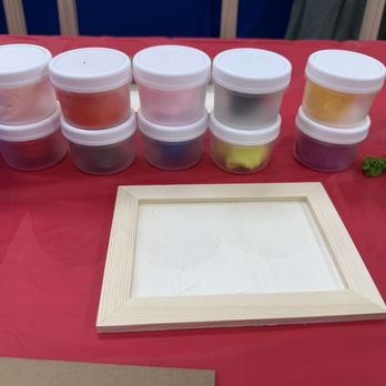 Crafty Tomato - Let the imagination begin! Air clay time!! - Small plastic containers with white lids, filled with colorful materials, are arranged on a red table, with a wooden frame and some green moss nearby. - Paint & Sip Near Me - Tempe, Arizona