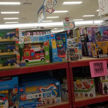 Ollie's Bargain Outlet - Toy shelves in a retail store are stocked with children's toys such as musical instruments, vehicles, and interactive playsets. The shelves are red and organized with different toy brands and types. A caricature of an elderly man with glasses and a big smile is hanging from the ceiling, positioned above the toy section. The store has bright fluorescent lighting and visible signage indicating sales and product categories. - Discount Store Near Me - Barkhamsted, Connecticut