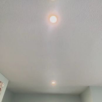 ProSpark Electric - A ceiling-mounted smoke detector with an illuminated indicator light is visible in a room with a window and a plant. - Security Systems Near Me