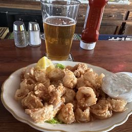 Vallemar Station Deep Fried Calamari - A plate of deep-fried calamari with lemon wedges and tartar sauce is placed on a wooden bar counter. Behind the plate, there is a glass of beer, a bottle of ketchup, and salt and pepper shakers. The background shows a bar area with wooden cabinets and bar equipment.