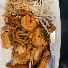 Pad Thai - A white rectangular plate contains Pad Thai noodles with shrimp, bean sprouts, and scrambled egg. The dish is topped with crushed peanuts and accompanied by a lime wedge. A metal fork and spoon are placed on the right side of the plate. The presentation suggests a typical Thai noodle dish served at a restaurant.