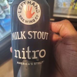 A person is holding a black can of Milk Stout Nitro from Left Hand Brewing Co., with the logo and product name clearly visible. The background shows a refrigerator with various beverage cans, indicating a retail or convenience store setting.