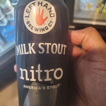 The Beer Keep - A person is holding a black can of Milk Stout Nitro from Left Hand Brewing Co., with the logo and product name clearly visible. The background shows a refrigerator with various beverage cans, indicating a retail or convenience store setting. - Beer Bar Near Me - Buffalo, New York