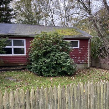 Vaz Construction - Before - A red wooden house with a moss-covered roof and small rectangular windows, surrounded by trees and a large bush in the yard, enclosed by a wooden fence. - Roofing Near Me - New Bedford, Massachusetts