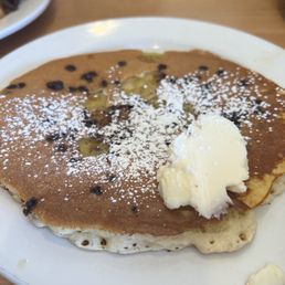 Pancakes - A close-up of a chocolate chip pancake on a white plate, topped with powdered sugar and a dollop of butter. The pancake appears thick and fluffy, with visible chocolate chips. The setting suggests a casual dining environment, likely at a breakfast and lunch spot known for creative dishes and fresh ingredients.