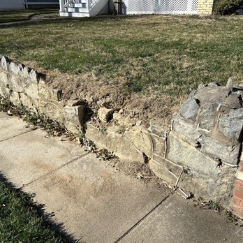 A&E Renovation - Old wall - The image depicts a stone and brick retaining wall adjacent to a concrete sidewalk in a residential setting. The stone section of the wall is visibly damaged, with some stones dislodged or broken, and the mortar appears eroded. The brick section on the right remains intact. The grassy area behind the wall is slightly elevated, and a house with stairs and a white railing is visible in the background. The scene suggests structural deterioration of the wall. - General Contractors Near Me - Falls Churs, Virginia