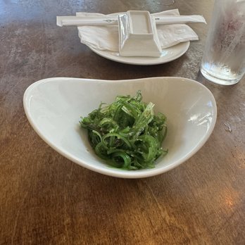 Yuzu Sushi - Seaweed Salad - A ceramic bowl filled with seaweed salad is placed on a wooden table. In the background, there is a glass of water with condensation and a plate with chopsticks wrapped in a napkin, along with a small rectangular dish. The setting appears to be a restaurant with natural lighting. - Japanese Near Me - Chamblee, Georgia