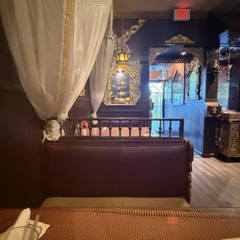 Ploy Thai - Entrance - The image shows a restaurant interior with a private booth separated by white curtains tied back. The booth has dark wood paneling and a cushioned seat. The background includes decorative wall art, ornate frames, and traditional-style lighting fixtures. The setting appears to be cozy and decorated with cultural elements, with a glimpse of the entrance and an exit sign visible. - Thai Near Me - Brandon, Florida