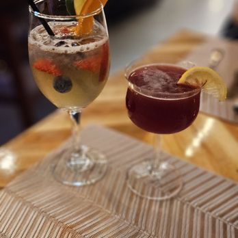 TapaVino - Two cocktails with fruit garnishes are placed on a wooden table in a restaurant setting, with a blurred background. - Modern European Near Me - Worcester, Massachusetts