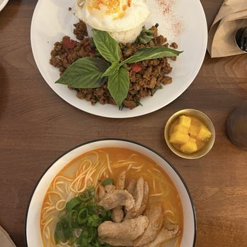 Momo Valley - Pad krapow gai and thukpa - The image shows a white plate with rice topped by a fried egg and garnished with fresh basil leaves, served with seasoned ground meat and vegetables. Next to it is a bowl of noodle soup containing chicken pieces, green onions, and thin noodles in a clear broth. A small dish of diced mango is also present on the wooden table, suggesting a combination of Asian-inspired dishes. - Himalayan/Nepalese Near Me - Beacon, New York