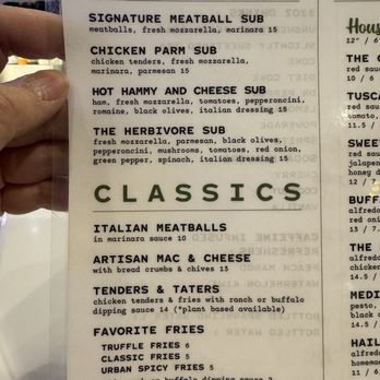Urban Pine Eatery - Classics - A printed menu displays sections for subs and classic dishes, including signature meatball, chicken parm, hot hammy and cheese, and herbivore subs, with descriptions and prices. The classics section offers Italian meatballs, artisan mac & cheese, tenders & taters, and various fries. A hand holds the menu, and a transparent protective cover partially covers the left side of the menu. - Pizza Near Me - Flagstaff, Arizona
