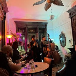 A live jazz performance takes place in a small, vintage-style room with ornate wall moldings and a large mirror. The scene is illuminated by colorful ambient lighting, including pink and blue hues. The band includes a double bass player and a saxophonist, with a vocalist standing between them. Audience members are seated at tables, enjoying the music, with drinks and glasses visible on the table. The ceiling features a decorative ceiling fan with large leaves.