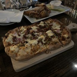 Goat Pistachio Flatbread - A rectangular pizza with melted cheese, chopped onions, and assorted toppings is served on a wooden board. The background shows a dark wooden table with glasses, plates, and a plate of fried chicken wings with dipping sauce. The setting appears to be a restaurant with a casual dining atmosphere.