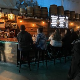 A group of people are seated at a bar inside a brewery, with barrels on the wall and a digital menu screen.