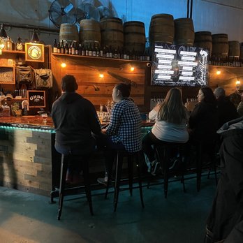 Ghost Hawk Brewing - A group of people are seated at a bar inside a brewery, with barrels on the wall and a digital menu screen. - Breweries Near Me - Clifton, New Jersey