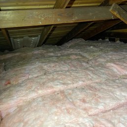 The image shows an attic space with visible wooden rafters and beams. The floor is covered with pink fiberglass insulation, which is used for thermal insulation in residential buildings. The insulation appears to be spread across the attic floor, and the space is enclosed by the roof structure. There are no other objects or features visible in the attic.