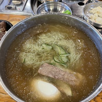 Jongro BBQ Flushing - Cold Green Tea Noodles - A bowl of cold green tea noodles with cucumber slices, a beef piece, and an egg in broth, served at a Korean restaurant. - Korean Near Me - Flushing, New York