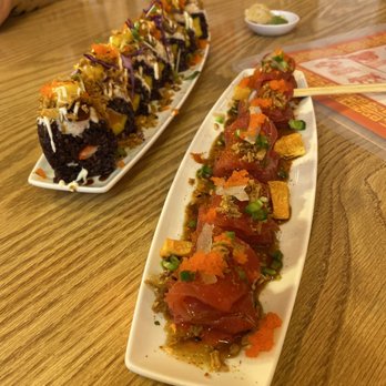 Dragon Sushi - Spicy citrus tuna & French Kiss Roll - The image shows two white, elongated plates of sushi on a wooden table. The front plate contains sushi with slices of raw fish, garnished with chopped green onions, orange fish roe, and sauce. The back plate features sushi rolls topped with colorful ingredients such as shredded vegetables, sauces, and possibly tempura flakes. In the background, there are small bowls, a paper menu, and a person wearing a polka-dot shirt partially visible. - Sushi Bars Near Me - Albuquerque, New Mexico