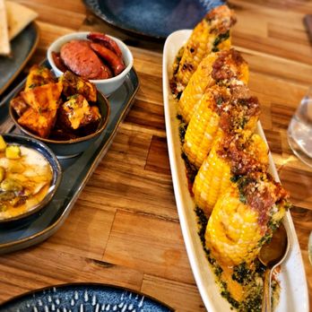 Oso - A long white platter holds several pieces of seasoned corn on the cob, garnished with herbs and cheese, on a wooden table. - Spanish Near Me - Silverton, Oregon