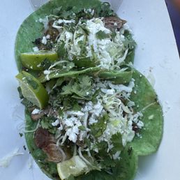 Green Butcher - Two tacos are presented on a paper tray, each with a green corn tortilla base topped with shredded cheese, cilantro, and pieces of meat. Lime wedges are placed beside the tacos for added flavor. The presentation suggests a casual dining setting, likely from a Mexican restaurant or food stand.