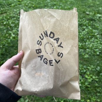 Sunday Bagels - A person holds a brown paper bag from Sunday Bagels, with the logo visible, outdoors on a grassy area. - Bagels Near Me - Albuquerque, New Mexico