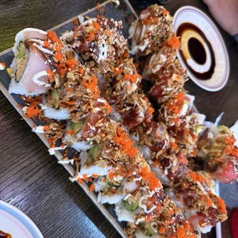 Dragon Sushi - Snow mountain, amazing, and another roll I can't remember but was delicious - A large sushi roll platter features multiple pieces of sushi topped with fish, orange fish roe, drizzled sauces, and crunchy toppings. The platter is placed on a dark wooden table, accompanied by soy sauce, a small bowl of sauce, and a bottle of soy or similar condiment. The presentation includes colorful ingredients and garnishes, with a hand visible in the background. - Sushi Bars Near Me - Albuquerque, New Mexico