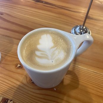 Southpine Bakery - Vanilla Latte - A latte with leaf-shaped foam art in a white ceramic mug is placed on a wooden table, with a metal straw in the background. - Coffee & Tea Near Me - Eugene, Oregon
