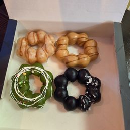 Strawberry Mochi Donut - A white box holds four donuts with different appearances. One glazed donut has a light caramel color with a drizzle of caramel sauce. Another donut is coated in dark chocolate with white salt crystals on top. A third donut is decorated with green icing, white icing drizzle, and colorful sprinkles, resembling a Christmas-themed treat. The fourth donut is black with a shiny glaze and a few salt crystals.