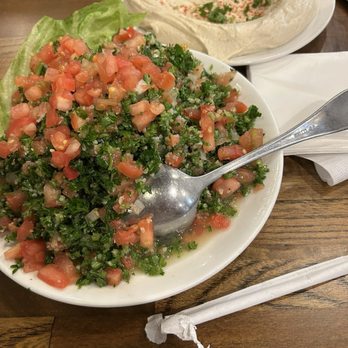 Al-Ameer - Tabouli & Hummus - A bowl of tabouli salad containing chopped tomatoes, parsley, and bulgur wheat, with a lettuce leaf on the side, placed on a wooden table. - Middle Eastern Near Me - Dearborn, Michigan