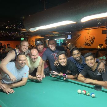 Atomic Billiards - Nine men are gathered around a pool table in a bar with orange walls, some leaning on the table and others standing behind. The bar has dim lighting with string lights and a large ventilation duct overhead. The background shows other patrons, pool tables, and a wall decoration of a lion. The group appears to be enjoying a social gathering, with some smiling and others engaged in the game. - Pool Halls Near Me - Washington, DC, District of Columbia