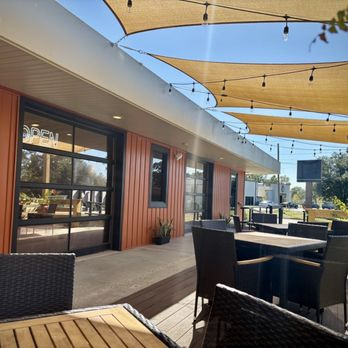 Empa Taco - Terrace seating at Empa Taco in Covington - An outdoor patio with tables and chairs, shaded by fabric canopies, is adjacent to a building with a neon 'OPEN' sign. - Latin American Near Me - Covington, Louisiana