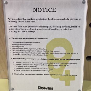 Rowan The Shoppes at Bethesda - 1.17.25 - A printed notice sign with safety procedures and risks related to skin-penetrating procedures, featuring a yellow skull graphic in the background. The sign is mounted on a wooden surface with four screws, and the bottom of the sign displays the business name 'Rowan.' The sign provides instructions for performing procedures safely and warns about potential health risks. - Jewelry Near Me - Bethesda, Maryland