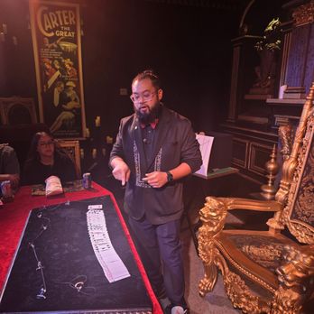 Mystique The Magic Experience - A man dressed in a black suit with a red bow tie performs a card trick at a table with a red cloth, surrounded by an audience in a dimly lit, ornate room. - Magicians Near Me - Mesa, Arizona