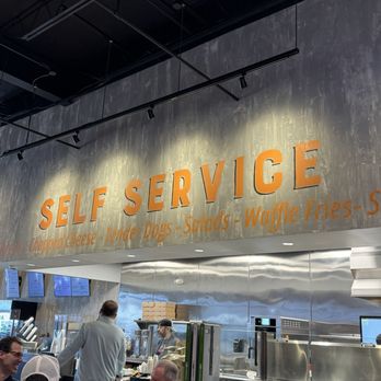 REV Burger - A self-service restaurant interior with a large sign reading 'SELF SERVICE' and menu items like burgers and salads, with customers seated and staff working behind the counter. - Burgers Near Me - Beverly, Massachusetts