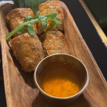 Siam Thai Eatery - Crispy Crab Roll (4) - Four spring rolls are arranged on a wooden platter with a small bowl of dipping sauce and garnishes, likely served at a Thai restaurant. - Thai Near Me - Chicago, Illinois