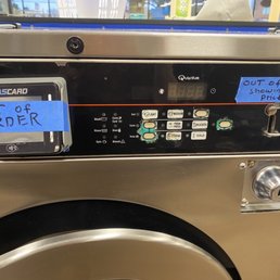 30+ machines have blue tape on them! - A stainless steel commercial washing machine features two blue sticky notes. The note on the card reader reads 'OUT OF ORDER,' and the note on the machine states 'OUT OF SERVICE showing wrong price.' The control panel includes various buttons and a digital display, with a key lock on the right side. The background shows a laundromat setting with windows and other equipment.