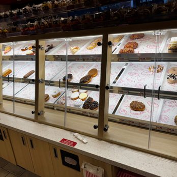 Kwik Trip - donuts - The image shows a glass display case filled with assorted donuts and baked goods, arranged on white paper liners. The case is part of a convenience store, with a wooden counter below and additional baked items stored on shelves above. Price tags are visible beneath the items, indicating their cost. The background includes a store interior with lighting and shelving. - Gas Stations Near Me - Roberts, Wisconsin